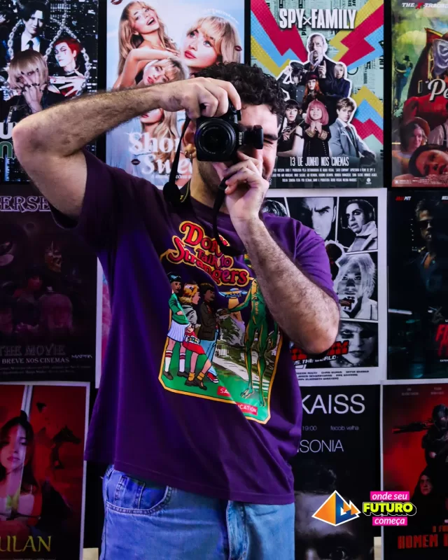 Homem fotografa com câmera em frente a pôsteres. UNIFAFIBE-13, ambiente criativo, camiseta estampada. Foco no futuro, artes visuais.
